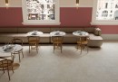 Gresowa podłogowa płytka imitująca beżowy kamień Saime Avalon Blanc Grip 60x60 8690102 na podłodze  restauracji. Jasna bezowa kanapa pod różową ścianą. Duże okna w białej ramie. Okrągłe stoliki na metalowej nodze. Drewniane krzesła z oparciem.