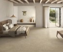 Gresowa podłogowa płytka imitująca beżowy beton Saime Avalon Beige Naturale 60x120 8690011 na podłodze przestronnej jasnej sypialni. Duże drzwi tarasowe ze szprosami w czarnej ramie. Drewniane łóżko z wiklinowym zagłówkiem.
