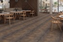 Płytka gresowa, podłogowa imitująca beton w brązowym kolorze, wykończenie powierzchni lappato, Netto Ceramika Cemento Rust Lappato 60x60 położona na podłodze w pomieszczeniu gdzie znajdują się drewniane jasne stoły oraz krzesła