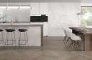 Płytka gresowa, podłogowa imitująca kamień w kolorze białym, wykończenie powierzchni satynowe, Pamesa Tresana Blanco Satin 90x90 położona na ścianie w kuchni gdzie znajduje się drewniana podłoga, stół oraz białe krzesła
