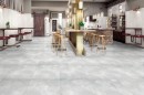 Płytka gresowa, podłogowa imitująca beton w szarym kolorze, wykończenie powierzchni lappato, Netto Ceramika Stardust Cemento Costa Del Grigio Lappato 60x60 położona na podłodze w pomieszczeniu gdzie znajdują się krzesła, stoliki oraz drewniane wykończenie