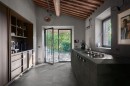 Nowoczesna kuchnia w rustykalnym stylu ma podłogę wyłożoną szarymi płytkami gresowymi Marazzi Mystone Pietra Ligure Cenere 75x75 cm MPKL, imitującymi kamień, wnętrze to charakteryzują ciemne meble, betonowy blat z płytą grzewczą i duże, przeszklone drzwi