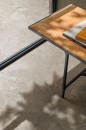 Bliskie ujęcie podłogi wyłożonej beżowymi płytkami gresowymi Marazzi Mystone Pietra Ligure Beige 75x75 cm MPKM, imitującymi kamień, na pierwszym planie widoczny jest metalowy stolik z miedzianym blatem i książkami, a na płytki pada cień liści