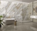 Gresowa podłogowa płytka imitująca biały marmur z wyraźnymi ciemnymi żyłami Saime Cave Silk Lucido 60x120-6mm T801977 na ścianie łazienki. Przyścienna okrągła wanna ze złotą podtynkową armaturą. Duża kabina typu Walk-in. Lustro w złotej ramie.