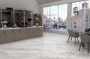 Płytka gresowa, podłogowa imitująca beton w szarym kolorze, wykończenie powierzchni matowe, Netto Ceramika Opal Grey Matt 60x120 położona na podłodze w pomieszczeniu gdzie znajdują się szare meble, krzesła oraz duże okno z widokiem na miasto