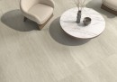 Płytka gresowa, podłogowa imitująca kamień w beżowym kolorze, wykończenie powierzchni matowe, Pamesa Dorset Taupe Matt 60x120 położona na podłodze w pomieszczeniu gdzie znajduje się jasny fotel oraz stolik kawowy