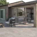 Płyta tarasowa imitująca kamień w kolorze beżowym, wykończenie powierzchni matowe, Pamesa Duplocem Taupe Matt 60x60 - 20mm położona na tarasie przed domem gdzie znajduje się fotel oraz drewniany stolik