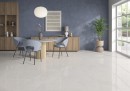 Płytka gresowa, podłogowa imitująca beton w niebieskim kolorze, wykończenie powierzchni matowe, Pamesa Eleganza Blu Matt 30x60 położona na ścianie w salonie gdzie znajdują się drewniane meble, stolik i krzesła oraz duże okno