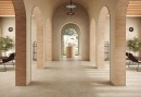 Przestronny hol wejściowy z beżową podłogą wyłożoną strukturalnymi płytkami Marazzi Mystone Tivoli Beige Strutturato 120x120 cm MQAT, płytki imitujące kamień tworzą spójną estetykę z łukami i ceglanymi elementami architektonicznymi