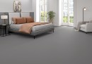 Płytka gresowa, podłogowa imitująca szary monokolorowy beton, wykończenie powierzchni matowe, Pamesa Essenza Grigio Matt 60x120 położona na podłodze w sypialni gdzie znajduje się łóżko, biały fotel oraz okna z widokiem na miasto