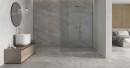 Płytka gresowa, podłogowa imitująca kamień w grafitowym kolorze, wykończenie powierzchni matowe, Pamesa Kenzo Merango Matt 60x120 położona na podłodze w łazience gdzie znajduje się prysznic, drewniana szafka z umywalką oraz wiszące lustro