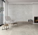 Płytka gresowa, podłogowa imitująca beton w szarym kolorze, wykończenie powierzchni matowe, Pamesa Midway Ash Matt 30x60 położona na ścianie w salonie gdzie znajduje się jasny fotel, stolik, kominek oraz okno z widokiem na ogród