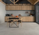 Płytka gresowa, podłogowa imitująca beton w szarym kolorze, wykończenie powierzchni matowe, Pamesa Mosel Sand Matt 60x60 położona na podłodze w kuchni gdzie znajduje się stół z krzesłami oraz drewniane meble kuchenne