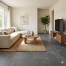 Płytka gresowa, podłogowa imitująca beton w grafitowym kolorze, wykończenie powierzchni matowe, Pamesa Mosel Grey Matt 90x90 położona na podłodze w salonie gdzie znajduje się jasna kanapa, drewniany stolik oraz szafka z telewizorem