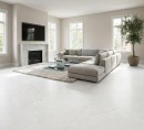 Płytka gresowa, podłogowa imitująca kamień w jasnym kolorze, wykończenie powierzchni matowe, Pamesa Natural Sense Blanco Matt 120x120 położona na podłodze w salonie gdzie znajduje się jasna kanapa, szklany stolik, kominek oraz okna z widokiem na ogród