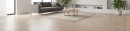 Nowoczesny salon z podłogą ułożoną w jodełkę francuską z jasnych, drewnopodobnych płytek Marazzi Nobilis Chevron Noce Sbiancato 11x54 cm MQ95, przestrzeń uzupełnia szara sofa, minimalistyczny drewniany stolik kawowy oraz duże okna