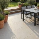 Płyta gresowa, tarasowa imitująca kamień w beżowym kolorze, wykończenie powierzchni matowe, Pamesa Pietra Di Rapolano Sand Matt 60x60 - 20mm położona na tarasie gdzie znajduje się ciemny stół, krzesła oraz rośliny ozdobne