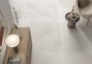 Płytka gresowa, podłogowa imitująca kamień w szarym kolorze, wykończenie powierzchni matowe, Pamesa Prince White Antic 120x120 położona na podłodze w salonie gdzie znajduje się jasny fotel, drewniana szafka oraz mały stolik
