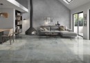 Płytka gresowa, podłogowa imitująca metal w zielonym kolorze, wykończenie powierzchni lappato, Pamesa Rusty Metal Moss Lappato 120x120 położona na podłodze w salonie gdzie znajduje się wypoczynek, kominek, stół z krzesłami oraz przejście na taras