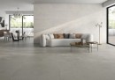 Płytka gresowa, podłogowa imitująca kamień w szarym kolorze, wykończenie powierzchni matowe, Pamesa Sospiro Gris Matt 120x120 położona na podłodze w salonie gdzie znajduje się jasny wypoczynek, stolik kawowy oraz duże okna