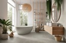 Jasna i przestronna łazienka wykończona wielkoformatowymi, szarymi płytkami Marazzi Room Grigio 100x100 cm MP6J, które imitują naturalny kamień na podłodze, nowoczesną aranżację dopełnia wolnostojąca wanna, drewniane meble oraz liczne rośliny