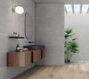 Płytka ścienna, dekoracyjna, imitująca beton w szarym odcieniu, wykończenie powierzchni matowe, ryflowane, Pamesa Dosso Greige Matt 40x120 położona na ścianie w łazience gdzie znajduje się drewniana szafka z białą umywalką oraz wiszące lustro i lampa