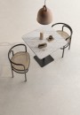Przytulna strefa kawiarniana z podłogą Ceracasa Coralina Ivory Matt 60x120. Jasny, matowy gres o wyglądzie kamienia tworzy naturalne tło dla marmurowego stolika i krzeseł z plecionką. Ciepły minimalizm w kolorach ziemi.