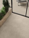 Wejście na taras z przeszkloną ścianą, długą drewnianą donicą z ozdobnymi roślinami oraz szarobeżowymi płytkami podłogowymi Caesar Slab2 Ash imitującymi kamień