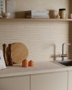 Nowoczesna kuchnia z pasem roboczym wykończonym beżowymi płytkami Marazzi Room Wall 3D Blaze Beige 20x60 cm MQ7J o strukturze 3D, trójwymiarowy, liniowy wzór kafli imitujących naturalny kamień stanowi stylowe tło dla jasnego blatu, drewnianej półki