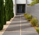 Nowoczesna ścieżka prowadząca do domu, ułożona z ciemnoszarych płyt tarasowych Marazzi Sentieri20 Nickel 60x60 - 20 mm MPZF rozdzielonych jasnym żwirem, minimalistyczną aranżację dopełniają wysokie, kolumnowe iglaki oraz nowoczesne, szare ogrodzenie