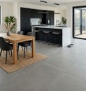 Nowoczesna kuchnia połączona z jadalnią, w której podłoga została wyłożona wielkoformatowymi płytkami Marazzi Slow Cold Salvia R10 60x60 MPEP, wnętrze uzupełniają czarne meble kuchenne, biała wyspa z marmurowym blatem oraz drewniany stół