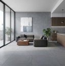 Nowoczesne, otwarte wnętrze łączące salon z kuchnią, w którym na podłodze i ścianie zastosowano duże, szare płytki Marazzi Slow Cold Piombo R10 60x60 cm MPEQ imitujące beton, pomieszczenie z oknami tarasowymi jest umeblowane grafitową sofą