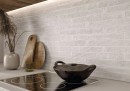 Płytka ścienna, cegiełka w białym kolorze, wykończenie powierzchni w połysku, Bestile Makalu Blanco Polished 5x50 położona na ścianie w kuchni gdzie znajduje się płyta indukcyjna oraz drewniane deski