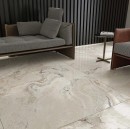 Płytka gresowa, podłogowa imitująca kamień w beżowym odcieniu, wykończenie powierzchni rustykalne w połysku, Ecoceramic Candy Blanco Antic Pulido 120x120 płytka położona na podłodze w salonie gdzie znajduje się szara kanapa oraz fotel