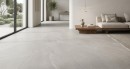Płytka gresowa, podłogowa imitująca kamień w szarym kolorze, wykończenie powierzchni lappato, Emigres Terra Gris Lappato 60x120 położona na podłodze w salonie gdzie znajduje się jasna kanapa oraz duże okna z widokiem na ogród