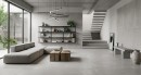 Nowoczesny salon z podłogą i elementami ścian wyłożonymi wielkoformatowymi, szarymi płytkami Marazzi Slow Cold Cemento 60x120 MPE9 imitującymi surowy beton, wnętrze uzupełnia nowoczesna sofa, drewniane stoliki i wiszący regał