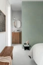 Sypialnia połączona z łazienką, w której zastosowano jasne płytki Marazzi Slow Wall Gesso 40x120 (MPC5). Minimalistyczne wnętrze z szałwiową ścianą, drewnianymi szafkami i okrągłym lustrem podkreśla subtelny, betonowy efekt płytek na podłodze.