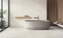 Gres Blooming Stone Alabaster White 60x120 w minimalistycznej łazience. Jasne płytki na podłodze i ścianie kontrastują z kamienną wanną wolnostojącą, drewnianymi lamelami oraz czarną armaturą. Przeszklone wyjście na taras.