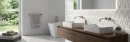 Jasna łazienka z płytkami Marazzi Slow Wall 3D Hay Gesso 40x120 MPC9. Biała ściana o trójwymiarowym, geometrycznym wzorze zestawiona z drewnianą szafką, dwiema umywalkami i wolnostojącą wanną. Nowoczesny, minimalistyczny design z widokiem na ogród.