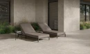 Gres Blooming Stone Sunny Beige 120x120 na nowoczesnym tarasie. Ciepłe, beżowe płyty pokrywają podłogę i elewację budynku. Dwa brązowe leżaki, mały stolik oraz ozdobne trawy tworzą luksusową strefę wypoczynku w naturalnym stylu.