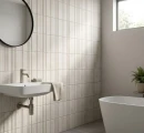 Nowoczesna łazienka ze ścianą wyłożoną strukturalnymi płytkami Marazzi ArtCraft Pomice Struttura 3D Bande 20x20 MGUD. Widoczna podwieszana umywalka, okrągłe lustro, fragment wanny oraz okno z widokiem na zieleń i wpadającym światłem słonecznym.