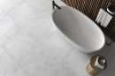 Gres Emigres America Blanco 80x80 na podłodze nowoczesnej łazienki. Jasne, betonopodobne płyty tworzą minimalistyczne tło dla białej wanny wolnostojącej i drewnianych lameli. Czarna armatura oraz surowy charakteru.