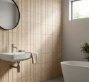 Łazienka ze ścianą w ciepłym beżu, wyłożoną strukturalnymi płytkami Marazzi ArtCraft Calce Struttura 3D Bande 20x20 cm MGUF. Widoczna biała umywalka, okrągłe lustro i wanna na tle pionowych, trójwymiarowych żłobień płytek w piaskowym odcieniu.