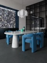 Nowoczesna, modernistyczna aranżacja jadalni. Z jednej strony surowy, grafitowy beton LaFaenza Party ALGAR6 120DG RM 120x120 na podłodze a z drugiej nowoczesne, niebieskie, zaokrąglone krzesła przy stole z jasnego marmuru.