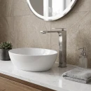 Wysoka bateria umywalkowa w nowoczesnym designie Noken Ibiza 100272816 w klasycznym chromowanym wykończeniu na marmurowym blacie połączonym z drewnianą szafką i białą owalną umywalką wolnostojącą.