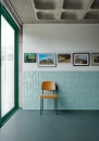Nowoczesne wnętrze z jasnoniebieskimi płytkami ściennymi Marazzi Luz Azzurro Polvere Lux 5,3x30 cm MFLS, drewnianym krzesłem i galerią obrazów na tle betonowego sufitu i dużego przeszklenia.