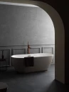Noken Square 100281868- wolnostojąca bateria wannowa o geometrycznym designie w kolorze miedzi szczotkowanej ze słuchawką i wężem w komplecie ustawiona przy wolnostojącej białej wannie. Szary beton na ścianach oraz podłodze.