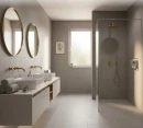 Nowoczesna łazienka wyłożona szarymi, technicznymi płytkami gresowymi Marazzi SistemT - Cromie Medio 30x30 cm MRJM. Płytki pokrywają podłogę i ściany, tworząc tło dla białej szafki z dwoma umywalkami, złotą armaturą i prysznicem ze złotą deszczownicą.
