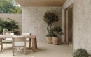 Fap Ceramiche Materia Pura Trama Beige R11 60x120- Gresowa płytka imitująca beżowy beton o strukturalnej dekoracyjnej powierzchni na podłodze zewnętrznego tarasu. Ściany pokryte gresową płytką imitującą lastryko. Drewniany stół tarasowy i krzesła.