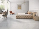 Ceracasa Davos Ice Polished 120x120- gresowa płytka imitująca jasny szary kamień z ciemnymi przebarwieniami na podłodze sypialni. Bezowe łóżko z oparciem, drewniana szafka nocna, drewniany stół. Biała boazeria dekoracyjna na ścianie.